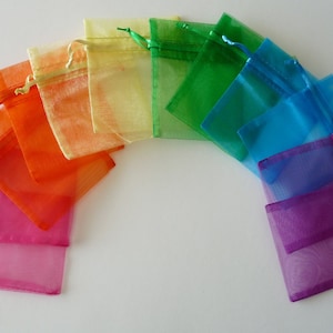 May include: A collection of colorful organza gift bags arranged in a rainbow arc. The bags are in shades of pink, orange, yellow, green, blue, and purple, each with a matching drawstring closure. The bags are semi-transparent.