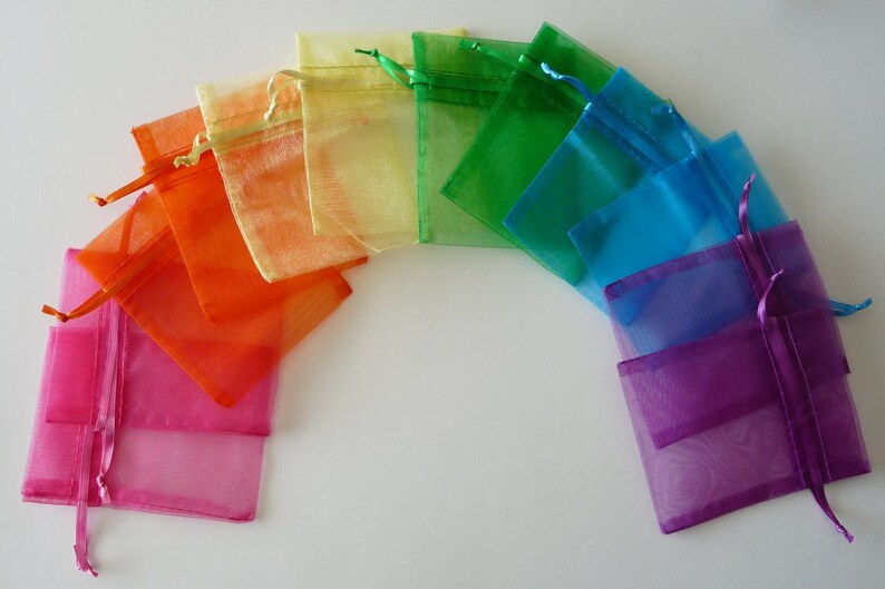 May include: A collection of colorful organza gift bags arranged in a rainbow arc. The bags are in shades of pink, orange, yellow, green, blue, and purple, each with a matching drawstring closure. The bags are semi-transparent.