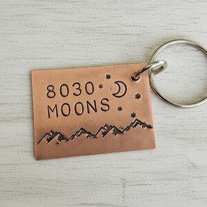 May include: A copper-colored rectangular keychain with the text "8030 MOONS" and a mountain range design. The keychain also features a crescent moon, stars, and a silver-colored key ring.