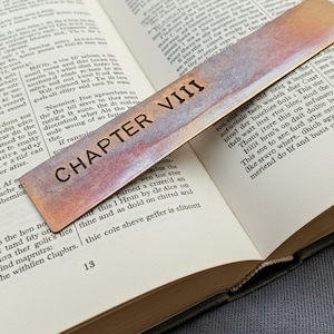 Könnte beinhalten: Ein rechteckiges Lesezeichen aus Metall mit einer verwitterten Kupferoberfläche und dunkler Patina. Der Text "CHAPTER VIII" ist in einer fetten, serifenlosen Schrift eingraviert. Das Lesezeichen liegt diagonal auf einem offenen Buch.