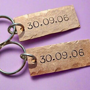 May include: Two copper-coloured rectangular keychains, each engraved with the date "30.09.06" in black. They are attached to silver key rings. The background is a solid purple colour.