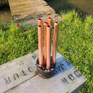 May include: A set of six copper pipes arranged in a circle and mounted on a wooden base. The pipes are all the same size and have a smooth, polished finish.