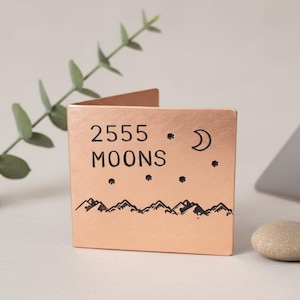 Könnte beinhalten: Ein kupferfarbener, quadratischer Metallkartenhalter mit dem Text "2555 MOONS" und einem Bergketten-Design. Das Design umfasst einen Halbmond und Sterne. Der Kartenhalter wird auf einer weißen Oberfläche präsentiert.