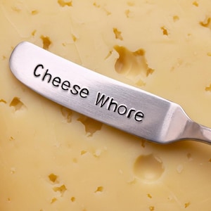 Könnte beinhalten: Ein Käsemesser aus Edelstahl mit den Worten "Cheese Whore" auf der Klinge liegt auf einem Block gelben Käses. Der Käse hat eine strukturierte Oberfläche mit kleinen Löchern.