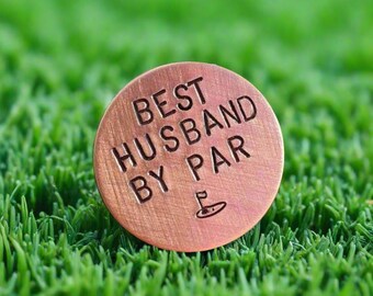 May include: A copper-colored golf ball marker with the text "BEST HUSBAND BY PAR" engraved on it. The marker also features a small golf flag icon.