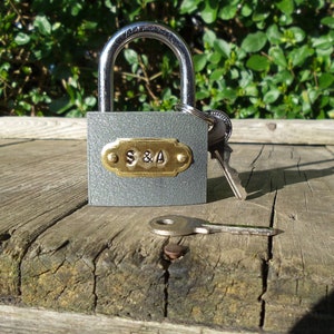May include: A gray padlock with a brass plate engraved with "S&A" and two keys. The padlock is sitting on a weathered wooden surface.