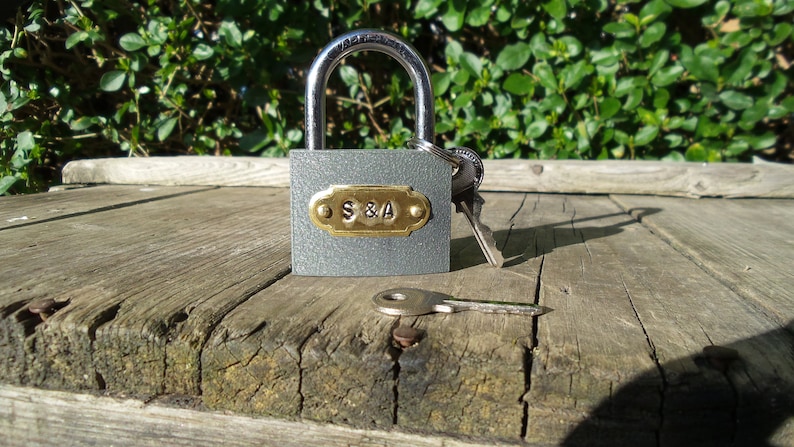 May include: A gray padlock with a brass plate engraved with "S&A" and two keys. The padlock is sitting on a weathered wooden surface.