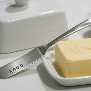 Op de afbeelding: Een roestvrijstalen botermes met het woord "Knob" op het lemmet ligt naast een blokje boter op een wit bord. Een witte boterdoos en een klein schaaltje boter staan ook op de foto.