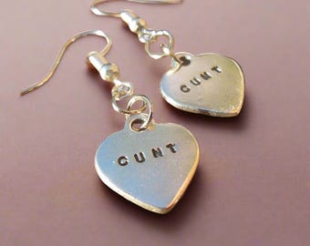 Puede incluir: Un par de pendientes plateados en forma de corazón. Cada corazón tiene la palabra "CUNT" estampada. Los pendientes tienen un diseño de gancho simple.