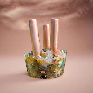 May include: A decorative, handcrafted holder with three copper-colored cylinders rising from a base of colorful, embedded stones and crystals. The base features a mix of green, yellow, and iridescent elements, creating a unique, artistic piece.