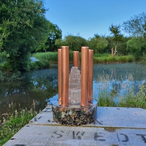 May include: A clear crystal tower with copper pipes surrounding it, set in a base of green and black stones. The tower is on a wooden surface with the word "SWEET" written on it.