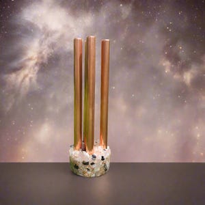 May include: A decorative copper candle holder with four vertical copper tubes. The base is made of a mix of small, colorful stones. The background features a starry, nebula-like design.