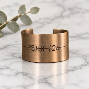 Op de afbeelding: Een brede armband van geborsteld bronskleurig metaal met een horizontale lijn in het midden gegraveerd. De cijfers 15/08/24 zijn in een eenvoudig, donker lettertype direct over de horizontale lijn gestempeld voor een datumontwerp.