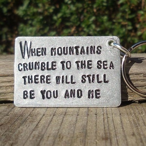 May include: A rectangular metal keychain with the quote "When mountains crumble to the sea there will still be you and me" engraved in black. The keychain has a silver key ring attached.