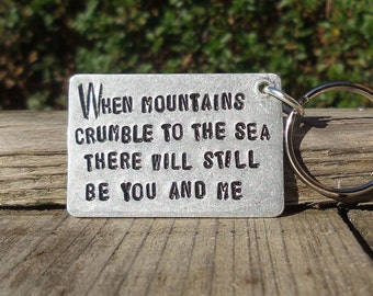 May include: A rectangular metal keyring with the quote "When mountains crumble to the sea there will still be you and me" engraved in black. The keyring has a silver key ring attached.