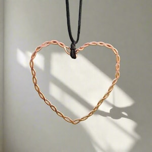 May include: A copper-colored heart pendant hangs from a black cord. The heart is crafted from twisted wire, creating a textured, open design. The pendant casts a shadow on a light-colored background, highlighting its shape and form.