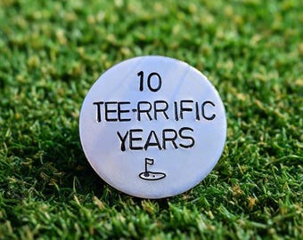 May include: A circular silver golf ball marker, featuring the inscription "10 TEE-RRIFIC YEARS" and a golf flag icon. The marker is positioned on a bed of green artificial grass.