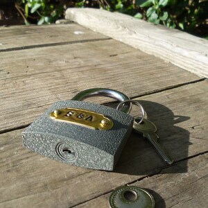 May include: A gray padlock with a brass plate engraved with the letters "S&A" and a silver key. The padlock is on a wooden surface.