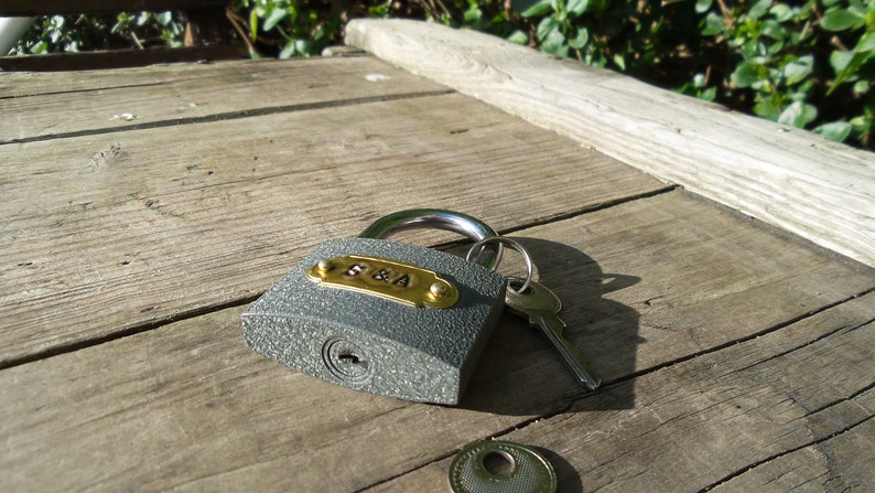May include: A gray padlock with a brass plate engraved with the letters "S&A" and a silver key. The padlock is on a wooden surface.