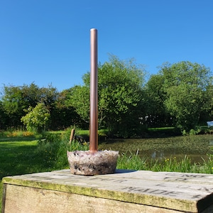 May include: A copper pipe inserted into a decorative concrete base with a pink and white marbled pattern. The base is sitting on a wooden surface.