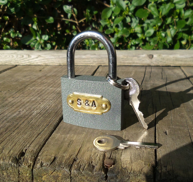 May include: A gray padlock with a silver shackle and a brass keyhole. The padlock has a brass plate with the inscription "S&A". The padlock is attached to a silver key with a brass keyhole.
