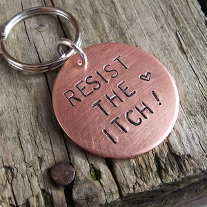 May include: A copper keyring with a silver key ring. The keyring has the text "Resist the itch!" engraved on it with a small heart symbol.