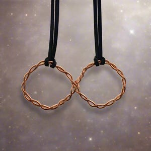 May include: A copper-colored infinity symbol pendant, crafted from twisted wire, hangs from a black cord. The pendant is suspended against a soft, speckled background, creating a minimalist and elegant design. The infinity symbol represents eternity and connection.
