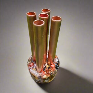 May include: A decorative vase featuring five copper tubes emerging from a base filled with colorful stones. The copper tubes are cylindrical and have a polished finish. The base is a rounded shape, showcasing a mosaic of various colored stones.