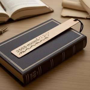 Könnte beinhalten: Ein Holzlesezeichen mit dem Text "2555 MOONS" und einem Bergketten-Design. Das Lesezeichen liegt auf einem geschlossenen, dunkelbraunen Buch mit goldenem Rand. Oben ist eine schwarze Schnur befestigt.