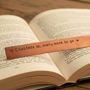 May include: A copper bookmark with the text "19 Chapters in, many more to go" and a heart symbol, resting on the open pages of a book. The bookmark is rectangular and has a slightly aged appearance.