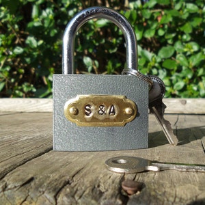 May include: A gray padlock with a brass plate engraved with the initials "S&A". The padlock is on a wooden surface with two keys.