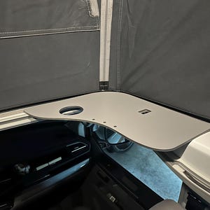 Könnte beinhalten: Ein hellgrauer, eckiger Tisch mit einem Getränkehalter und mehreren kleinen Löchern. Der Tisch ist in einem Fahrzeug montiert, mit einem dunkelgrauen Stoff darüber. Das Innere des Fahrzeugs ist darunter sichtbar.