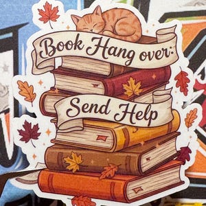 May include: A sticker featuring a stack of books in shades of brown and orange, with a sleeping orange tabby cat on top. The sticker has the text "Book Hangover: Send Help" on banners, with autumn leaves scattered around.
