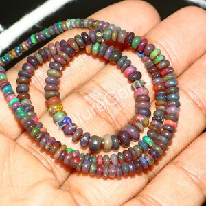 May include: A close-up of three strands of opal bead necklaces. The beads are small, oval-shaped, and display a variety of colors, including black, green, red, and purple. The necklaces are held in a person's hand.