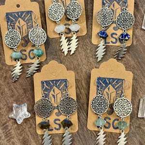 May include: Earrings with intricate silver filigree circles, gemstone beads, and lightning bolt charms. The earrings are displayed on brown tags with a blue logo. The gemstones include turquoise, white, blue, and green. The earrings are a bohemian style.
