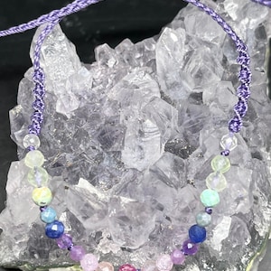 Pode incluir: Um bracelete de pedras preciosas arco-íris com cordão roxo. O bracelete apresenta uma variedade de pedras preciosas redondas e coloridas, incluindo verde, azul, roxo, rosa e vermelho.
