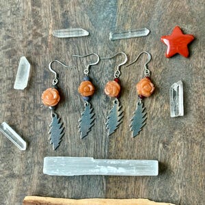 May include: Three pairs of earrings with orange rose-shaped beads and silver leaf charms. The earrings are displayed with clear quartz crystals, a red star, a selenite wand, and a piece of wood on a wooden surface.