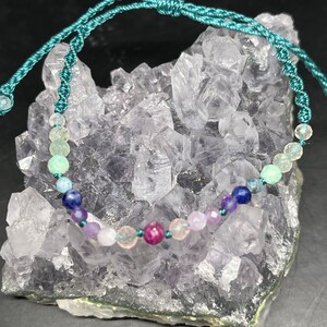 Pode incluir: Um bracelete de corda turquesa com uma variedade de contas de pedras preciosas, incluindo verde, azul, roxo, rosa e vermelho.