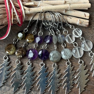 May include: A collection of handmade earrings with silver tree charms and gemstone beads in various colors, including green, purple, and clear. The earrings are displayed on a wooden surface with a bundle of sticks and a star-shaped crystal.