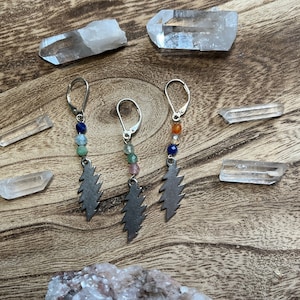 May include: Three pairs of earrings with a silver lightning bolt charm and colorful gemstone beads. The earrings are displayed on a wooden surface with clear quartz crystals.