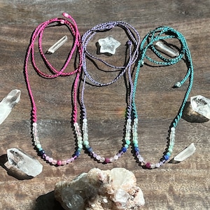 May include: Three colorful gemstone necklaces with a gradient of colors, including pink, purple, blue, and green. The necklaces are made with small, round beads and are strung on a braided cord. The necklaces are displayed on a wooden surface with clear quartz crystals.