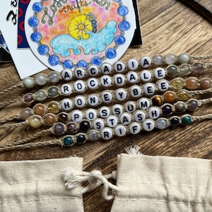May include: Seven beaded bracelets with different colored stones and white letter beads spelling out the words "ARCADIA", "ROCKDALE", "HONEYBEE", "POSSUM", "STASH", and "RIFT". The bracelets are on a wooden surface with a small white bag.