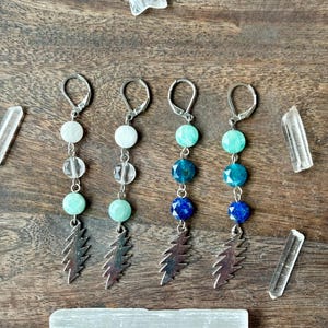 May include: Four pairs of dangle earrings with silver-tone lightning bolt charms. Each earring features a combination of round, colorful beads in shades of white, green, and blue. The earrings are displayed on a wooden surface with crystals.