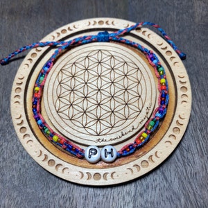 May include: A colorful beaded bracelet with the letters "PH" on white beads. The bracelet is on a wooden coaster with a flower of life design and the text "the weekend mystic".