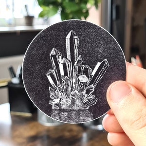 May include: A round, black sticker featuring a detailed white illustration of a cluster of crystals. The crystals are depicted with sharp, angular facets, creating a striking contrast against the dark background. The sticker is held by a hand.