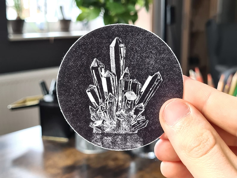 May include: A round, black sticker featuring a detailed white illustration of a cluster of crystals. The crystals are depicted with sharp, angular facets, creating a striking contrast against the dark background. The sticker is held by a hand.