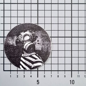 May include: A black and white circular sticker featuring a person wearing a gas mask and a striped shirt. The design is a detailed illustration with a vintage aesthetic. The sticker is placed on a grid background.
