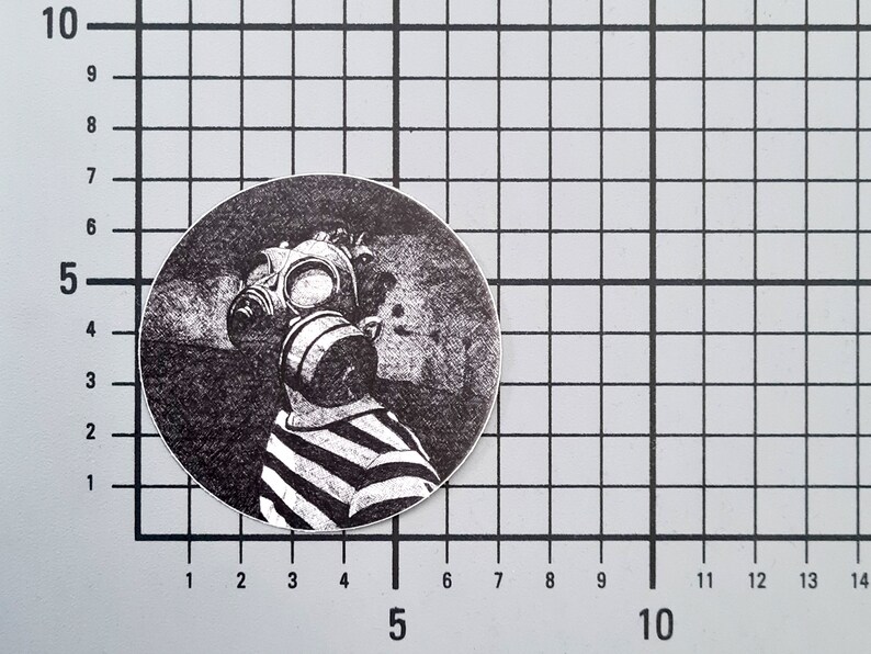 May include: A black and white circular sticker featuring a person wearing a gas mask and a striped shirt. The design is a detailed illustration with a vintage aesthetic. The sticker is placed on a grid background.