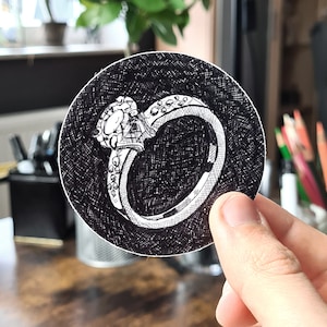 Könnte beinhalten: Eine Schwarzweißillustration eines Diamant-Verlobungsringes mit einem großen Mittelstück und kleineren Steinen um die Band. Die Illustration befindet sich auf einem runden Aufkleber.