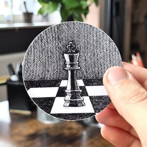 Puede incluir: Ilustración en blanco y negro de un rey de ajedrez sobre una pegatina redonda. El rey está centrado en un tablero de ajedrez, con un fondo texturizado. La pegatina está sujeta por una mano.